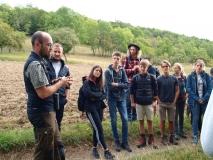 Eine Gruppe junger Erwachsener vor einem Acker und Wald schaut aufmerksam auf einen Exkursionsleiter