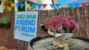 Ein Banner des MAB Jugendforums 2023 hängt an einem Zaun. Im Vordergrund ein Topf Heidekraut.