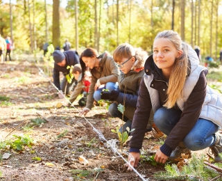 Eine Gruppe Jugenlicher kniet auf dem Waldboden und planzt Bäume. Ein Mädchen im Vordergrund des Bildes blickt lächelnd in die Kamera.