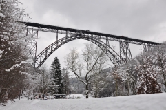 Aus der Perspektive der Wupperaue ist die stählerne und markant wirkende Bogenbrücke zu sehen. Es ist ein verschneiter, grauer Wintertag.