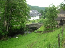 Das Foto zeigt ein typisches Kerbsohlental in der Ruhreifel mit Bach, Bachbegleitgehölzen, einer Steinbrücken und typischen Naturstein- und Fachwerkhäusern der Eifel.