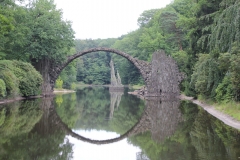 Es ist eine bogenförmige Basaltsäulen-Brücke am Rakotzsee im Kromlauer Park abgebildet. Die Brücke spiegelt sich eindrucksvoll auf der Wasserfläche des Sees.